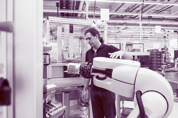 Auf dem Bild ist zu sehen, wie ein Mann den Arm eines Industrieroboters steuert. Der Mann hat lange dunkle Haare.