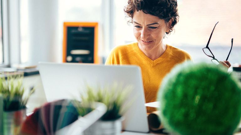 Eine Frau sitzt lächelnd vor einem Laptop, in der linken Hand hält sie eine Brille